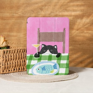 May include: A pink tablet case with a playful design of a black and white cat holding a cocktail. The cat is at a table with a green and white checked tablecloth, and a plate with a fish design.