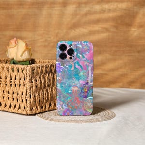 Pode incluir: Uma capa de telefone colorida com um design abstrato vibrante em tons de azul, rosa e roxo. A capa apresenta um recorte multicâmera e está sobre um porta-copos redondo e tecido. Uma cesta de vime com uma rosa está no fundo.