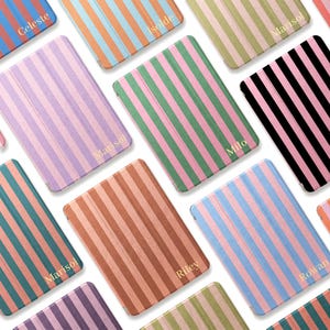 Puede incluir: Una colección de fundas rectangulares para tabletas con rayas verticales en varias combinaciones de colores, incluyendo rosa, verde, azul, naranja, negro y marrón. Cada funda está personalizada con un nombre en color dorado como Celeste, Isolde, Marisol, Milo, Riley o Rowan.