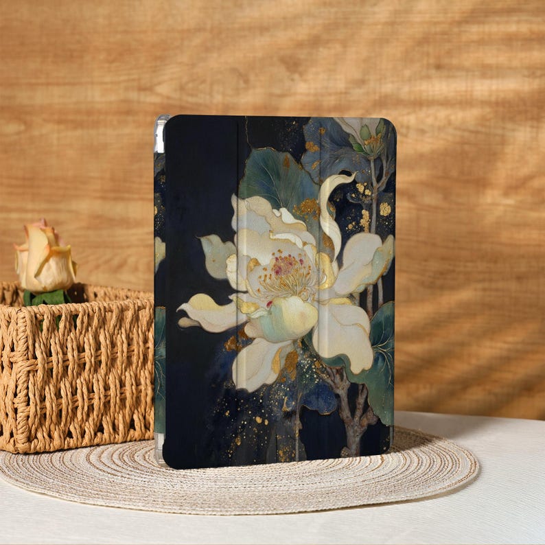 May include: A tablet case featuring a large, stylized white flower with gold accents and green leaves against a dark blue background. The floral design is reminiscent of a watercolor painting. The case is on a neutral-colored surface.