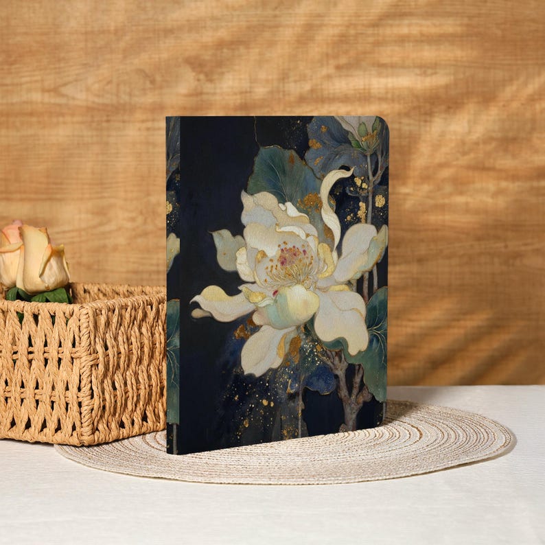 May include: A hardcover journal featuring a large white flower with yellow and gold accents against a dark blue background. The floral design includes green leaves and stems. The journal is displayed on a white surface.