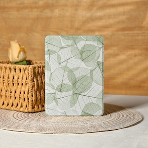 May include: A rectangular tablet case featuring a repeating pattern of translucent green leaves on a white background. The case is displayed on a woven placemat, with a wicker basket and a yellow rose in the background.