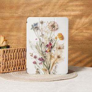 Pode incluir: Capa de tablet branca com design floral. A imagem mostra um buquê de flores secas em tons de roxo, amarelo e creme. A capa é retangular com cantos arredondados, e o arranjo floral está centrado na frente.
