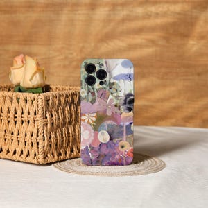 Op de afbeelding: Een smartphonehoesje met een bloemenontwerp in tinten paars, roze en groen. De hoes heeft een collage van bloemen en abstracte vormen. De telefoonhoes ligt op een ronde, geweven onderzetter.