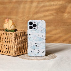 May include: A white smartphone case with a celestial design. It features light blue watercolor clouds, stars, and constellations. The case has a glossy finish and a black camera module. The design includes a crescent moon and a small figure on a crescent moon.