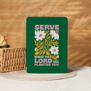 Peut inclure: Une coque de tablette verte avec l'inscription "SERVE WHERE THE LORD HAS PLANTED YOU" en blanc et rose, ornée de fleurs blanches et de feuilles jaunes. L'étui est posé sur une surface blanche.