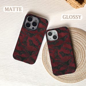May include: Two phone cases with a repeating pattern of black bats on a deep red background. One case is matte, the other is glossy. The cases are on a light-colored surface.