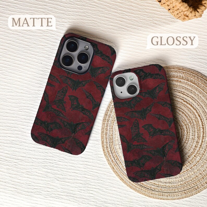 May include: Two phone cases with a repeating pattern of black bats on a deep red background. One case is matte, the other is glossy. The cases are on a light-colored surface.
