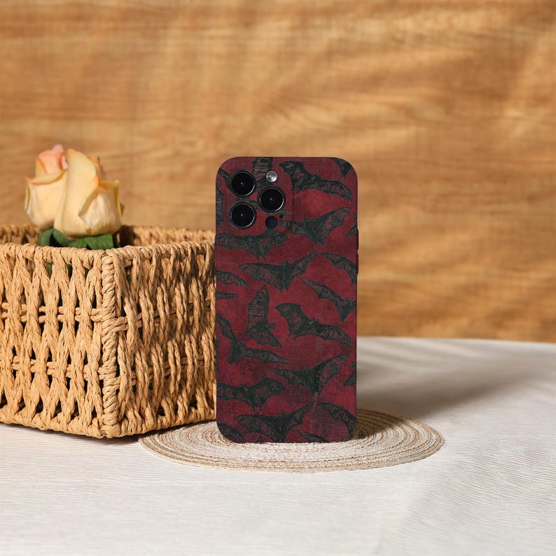 May include: A burgundy phone case featuring a repeating pattern of black bat silhouettes. The case has a glossy finish and is designed to fit a smartphone. The background includes a woven basket with a rose and a wooden surface.