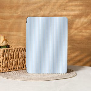 Puede incluir: Una funda rectangular para tableta con un patrón de rayas verticales en azul claro y blanco. La funda está sobre un salvamanteles de color neutro, con una cesta tejida y una rosa en el fondo. El fondo es una superficie de madera.