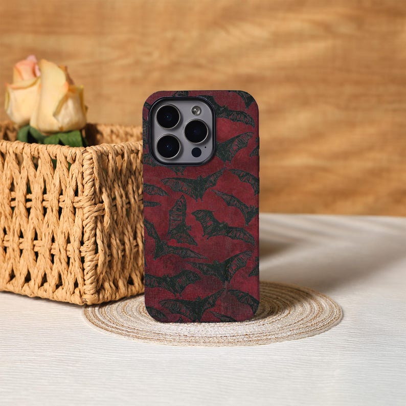 May include: A phone case with a dark red background and a repeating pattern of black bats. The case has a camera cutout and is displayed on a white surface with a woven basket and a rose in the background.