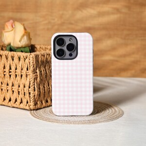 May include: A white and pink gingham patterned phone case. The phone case is standing upright on a round, woven placemat. A woven basket with a yellow rose is in the background.