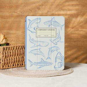 Puede incluir: Una cubierta de cuaderno de composición azul claro con ilustraciones de tiburones azules. La cubierta del cuaderno tiene el texto "COMPOSITION NOTEBOOK" y mide 19,05cm x 23,5cm.