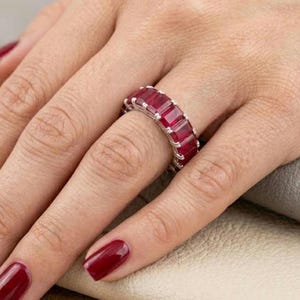 Peut inclure: Une bague en argent ornée d'une bande continue de pierres précieuses rectangulaires rouge foncé. La bague est portée au doigt, avec la main et une surface de couleur neutre visibles en arrière-plan. Les pierres sont espacées et serties dans une monture en argent.