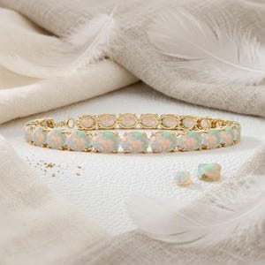 Puede incluir: Una pulsera de tono dorado con una fila continua de ópalos ovalados e iridiscentes engastados en garras de metal. Dos ópalos ovalados sueltos están colocados cerca de la pulsera, mostrando tonos blanco lechoso con destellos de color.