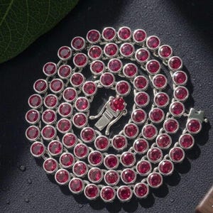 May include: A circular tennis necklace featuring numerous round, deep red gemstones set in polished silver-toned metal bezels. The necklace is arranged in a coiled spiral pattern and includes a secure, rectangular box clasp mechanism at the end.
