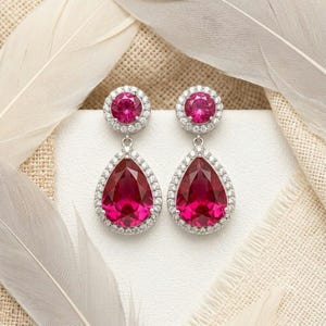 May include: A pair of drop earrings featuring a round, faceted, deep pink stone at the top, surrounded by a halo of small clear stones, connected to a larger, pear-shaped, deep pink stone at the bottom, also framed by a halo of clear stones.