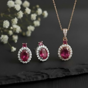 Op de afbeelding: Een sieradenset met een hangerketting en bijpassende oorbellen. De set bevat een gouden ketting met een robijn hanger en twee robijn oorbellen. Elk stuk is versierd met kleine, sprankelende witte edelstenen. De sieraden worden gepresenteerd op een zwart oppervlak.