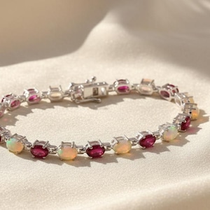 Op de afbeelding: Een zilveren armband met afwisselende ovale edelstenen. De armband toont levendige rode en iriserende opaalachtige stenen, vastgezet met een zilveren sluiting. De sieraden worden gepresenteerd op een lichtgekleurd, getextureerd oppervlak.