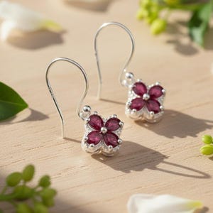 May include: A pair of silver drop earrings featuring a four-petaled flower design. Each earring is set with four marquise-cut deep red gemstones arranged in a clover shape, accented by small clear stones along the silver metal setting.
