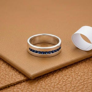 Puede incluir: Un anillo plateado con una fila de pequeñas piedras preciosas azul oscuro. El anillo está sobre una superficie de cuero marrón. Un rollo de papel blanco está en el fondo.