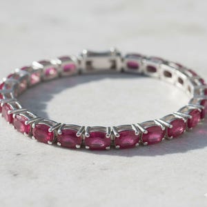 Puede incluir: Una pulsera de plata con una hilera de piedras preciosas rojas ovaladas. Las piedras están engarzadas juntas, creando una línea continua de color. La pulsera tiene un cierre seguro.