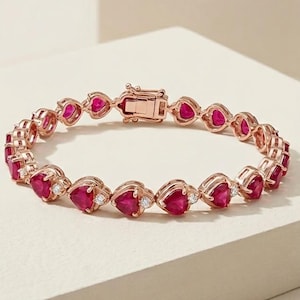 May include: Rose gold bracelet with heart-shaped red gemstones and small white accent stones. The bracelet has a secure clasp. The jewellery is displayed on a neutral surface. A piece of jewellery for special occasions.
