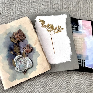 May include: Three handmade cards with botanical and abstract designs. One card features a dried flower arrangement with a wax seal. Another has a dried plant on white paper. The third card has a grid and watercolor design.