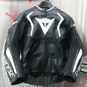 Può includere: Giacca da moto in pelle nera con dettagli bianchi e logo bianco sul petto. La giacca ha un colletto rialzato e strisce bianche sulle maniche. Il nome del marchio è visibile sul colletto e sulle maniche.