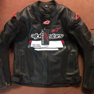 Puede incluir: Chaqueta de moto de cuero negro con detalles rojos y blancos. Presenta el logotipo "Alpinestars" en el pecho y las mangas. Incluye el texto "ONE GOAL ONE VISION".