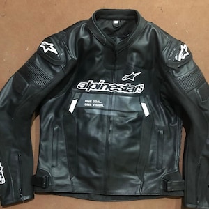 Op de afbeelding: Zwarte leren motorjas met witte Alpinestars-logo en accenten. Voorzien van een opstaande kraag, gewatteerde schouders en manchetten met rits. De tekst "ONE GOAL ONE VISION" staat op de voorkant.