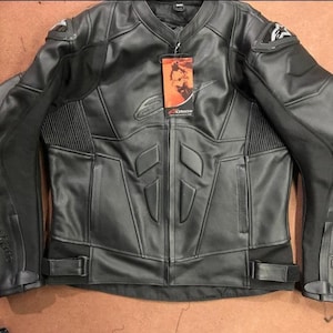 Può includere: Giacca da moto in pelle nera con collo alla coreana, chiusura con zip e spalle imbottite. La giacca presenta un'etichetta con un logo ed è progettata con pannelli rinforzati e dettagli a coste.