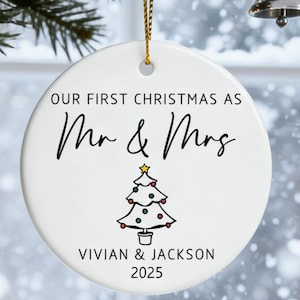 May include: A white ceramic Christmas ornament with the text "OUR FIRST CHRISTMAS AS Mr & Mrs" in black script. A small Christmas tree illustration is below the text, with the names "VIVIAN & JACKSON" and the year "2025" below the tree.
