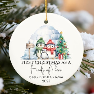 May include: A white ceramic Christmas ornament featuring a family of penguins in winter attire. The ornament has the text "First Christmas as a Family of Three" and the names "Dad + Sophia + Mom 2025".