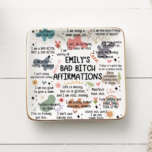 Könnte beinhalten: Ein rechteckiges Keramiktablett mit goldenem Rand, auf dem in Schwarz und Rosa "Emily's Bad Bitch Affirmations" steht. Das Tablett ist mit Affirmationen und farbenfrohen Grafiken, darunter Blumen und Sterne, verziert.