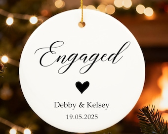 May include: A white, circular Christmas ornament with the word "Engaged" in script, a black heart, and the names Debby & Kelsey with the date 19.05.2025. It hangs from a gold string, with a Christmas tree and lights in the background.