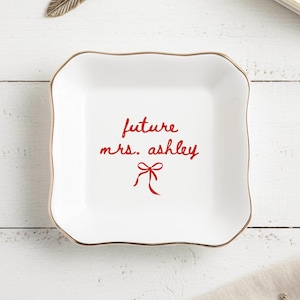 May include: A white ceramic trinket dish with a scalloped edge and gold trim. The dish features the red text "future mrs. ashley" with a bow graphic. Suitable for holding jewellery or small items.