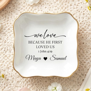 Puede incluir: Un plato de cerámica blanco con borde dorado, con la inscripción "we love BECAUSE HE FIRST LOVED US 1 John 4:19 Maya ♥ Samuel". El plato está rodeado de flores blancas y se encuentra sobre una superficie de encaje.
