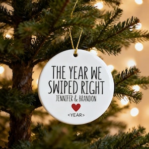 May include: Round white ceramic Christmas tree ornament with gold string. Text reads "THE YEAR WE SWIPED RIGHT", with names and a red heart. The ornament is on a Christmas tree. The year is indicated by "<YEAR>".