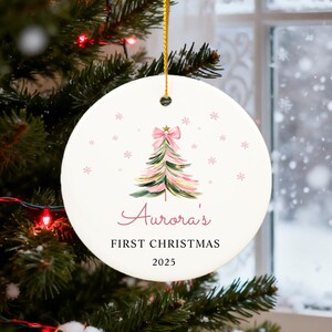 May include: A round, white Christmas ornament with a watercolor-style Christmas tree design in pink and green, topped with a pink bow. The text reads "Aurora's FIRST CHRISTMAS 2025". The ornament hangs from a gold string.
