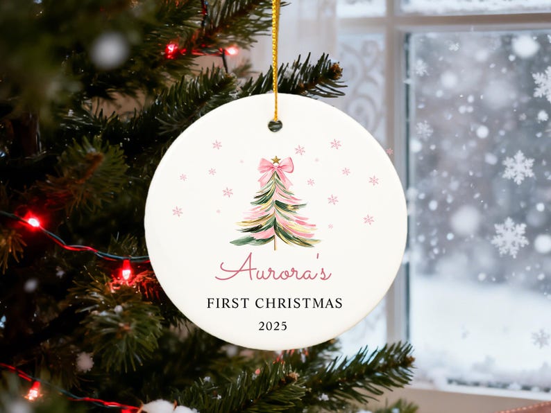 May include: A round, white Christmas ornament with a watercolor-style Christmas tree design in pink and green, topped with a pink bow. The text reads "Aurora's FIRST CHRISTMAS 2025". The ornament hangs from a gold string.