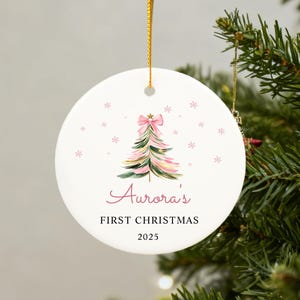 May include: A white ceramic Christmas ornament with a watercolor Christmas tree design in pink and green, topped with a pink bow. The ornament reads "Aurora's FIRST CHRISTMAS 2025" in pink script and black text. A gold string is attached for hanging.