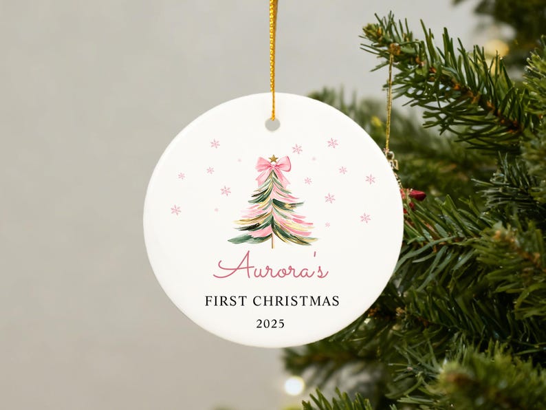 May include: A white ceramic Christmas ornament with a watercolor Christmas tree design in pink and green, topped with a pink bow. The ornament reads "Aurora's FIRST CHRISTMAS 2025" in pink script and black text. A gold string is attached for hanging.