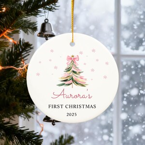 May include: A round Christmas ornament with a watercolor Christmas tree design in pink and green, topped with a pink bow. The text "Aurora's FIRST CHRISTMAS 2025" is written in a script font. The ornament hangs from a gold ribbon.