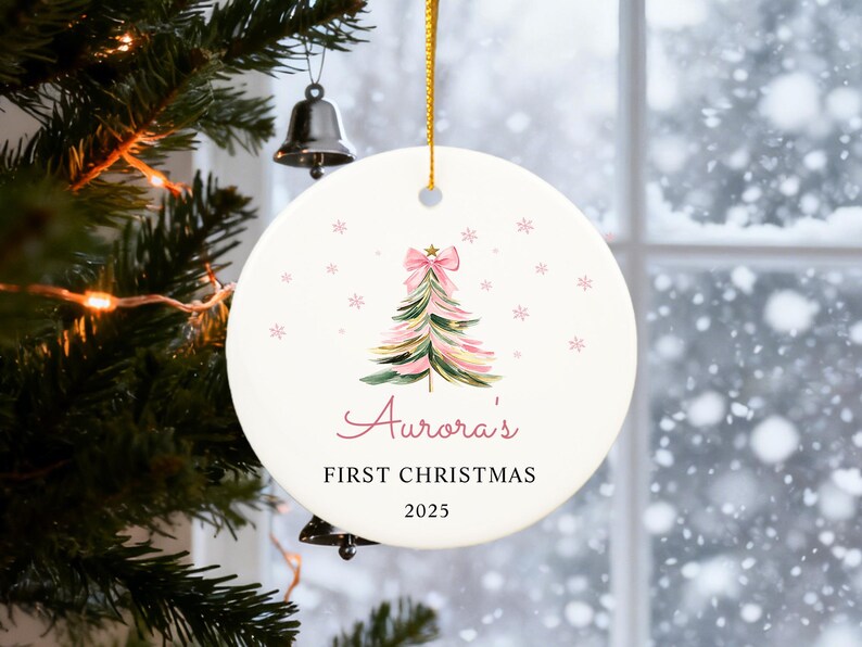 May include: A round Christmas ornament with a watercolor Christmas tree design in pink and green, topped with a pink bow. The text "Aurora's FIRST CHRISTMAS 2025" is written in a script font. The ornament hangs from a gold ribbon.