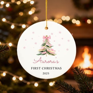 May include: A round, white Christmas ornament with a watercolor Christmas tree design, a pink bow, and snowflakes. The text "Aurora's FIRST CHRISTMAS 2025" is written in a script font. The ornament hangs from a gold string.
