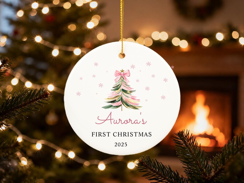 May include: A round, white Christmas ornament with a watercolor Christmas tree design, a pink bow, and snowflakes. The text "Aurora's FIRST CHRISTMAS 2025" is written in a script font. The ornament hangs from a gold string.