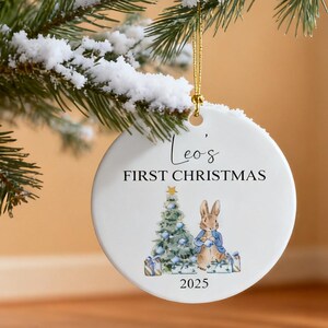 Puede incluir: Un adorno navideño de cerámica blanca con una cuerda dorada, con una ilustración de acuarela de un árbol de Navidad, un conejo y el texto "Leo's FIRST CHRISTMAS 2025". El adorno cuelga de una rama de pino nevada.