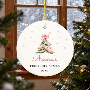 May include: A round Christmas ornament with a watercolor Christmas tree design. The tree is pink and green with a pink bow at the top. The text "Aurora's FIRST CHRISTMAS 2025" is written in pink script. A gold string is attached for hanging.