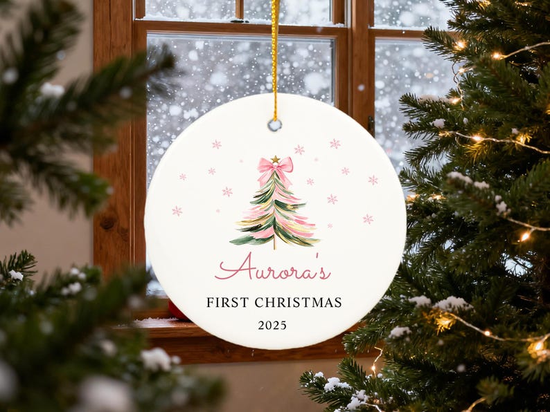 May include: A round Christmas ornament with a watercolor Christmas tree design. The tree is pink and green with a pink bow at the top. The text "Aurora's FIRST CHRISTMAS 2025" is written in pink script. A gold string is attached for hanging.